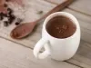 Imagem de uma xícara branca com chocolate quente e ao lado uma colher de pau pequena com chocolate em pó.